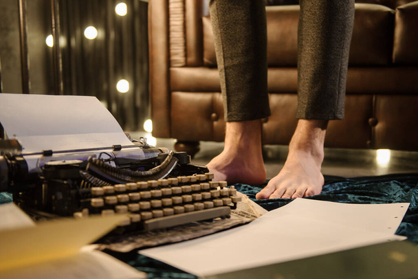 közelkép férfi lábairól egy régi írógéphez közel egy újságpapíron és sötétkék bársony szövet a padlón egy sötét szobában, izzókkal egy barna bőrkanapé mellett. - Fotó, kép