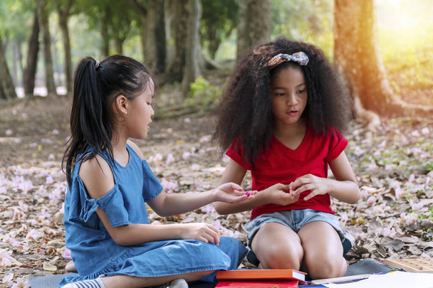 Cheerful two kid girl holding plasticine looking at something while sitting together in the park.Mixed race schoolchild with plasticine in her hands while playing with asian friend.Friendship concept. - Foto, Bild