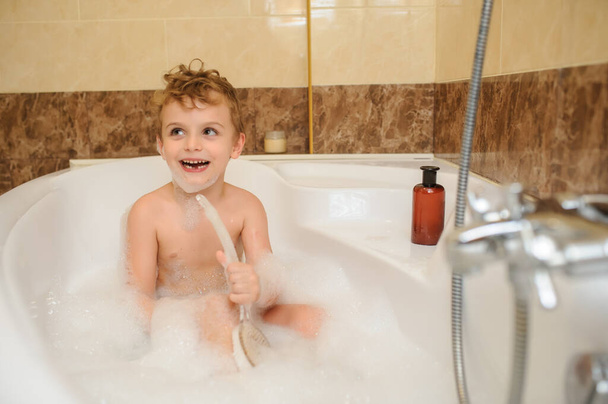 Μικρό αγόρι πλένει και παίζει στην μπανιέρα με αφρό και σαπουνόφουσκες - Φωτογραφία, εικόνα