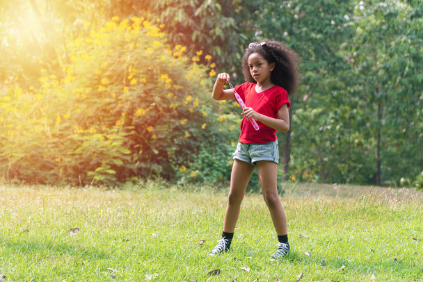 Mischlingskind mit lockigem Haar, das Seifenblasen hält, während es auf dem Rasen im öffentlichen Park spaziert. Kleine süße Mädchen positiv spielen Seifenblasen fühlen sich entspannt auf der Natur Hintergrund mit Kopierraum. - Foto, Bild