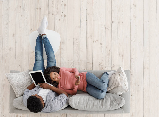 Weekend Leisure. Black Couple Relaxing On Couch With Digital Tablet And Smartphone - Photo, Image