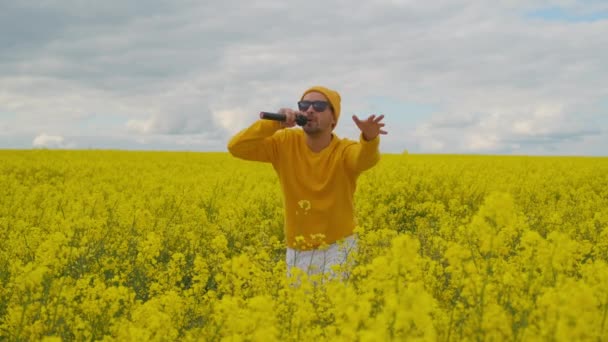 Nosząc okulary przeciwsłoneczne i pole żółtych kwiatów, męski solista w żółtych ubraniach tańczy i śpiewa energiczną piosenkę tańcząc i machając rękami. Kamera na żywo - Materiał filmowy, wideo