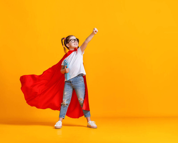 Koko kehon tyttö supersankari viitta hymyillen ja nostamalla nyrkki ylös ollessaan valmis koulun tutkimukset vastaan keltainen backdro - Valokuva, kuva