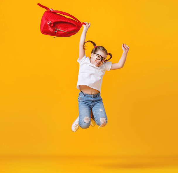Pełne ciało podekscytowany dziewczyna z czerwonym plecakiem uśmiecha się i skacze w górę podczas studiów w szkole przed żółtym backdro - Zdjęcie, obraz