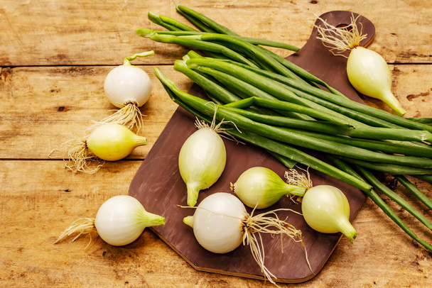 Fresh young onion and its greens. Traditional ingredient for cooking. Cutting board, old wooden background - Valokuva, kuva