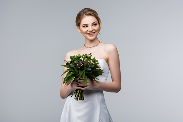 happy bride in white dress holding wedding flowers isolated on grey  - 写真・画像