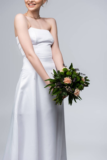 przycięty widok szczęśliwej panny młodej w białej sukience gospodarstwa kwiaty izolowane na szarości  - Zdjęcie, obraz