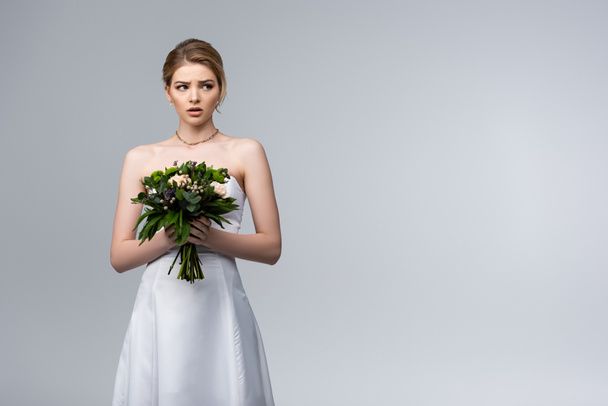 verrast bruid in witte trouwjurk met bloemen geïsoleerd op grijs  - Foto, afbeelding