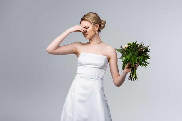 missmutige Braut im weißen Hochzeitskleid berührt Nase, während sie stinkende Blumen isoliert auf grau hält  - Foto, Bild