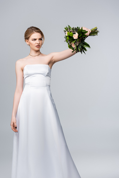 dissatisfied bride in white dress looking at wedding flowers isolated on grey  - Photo, Image