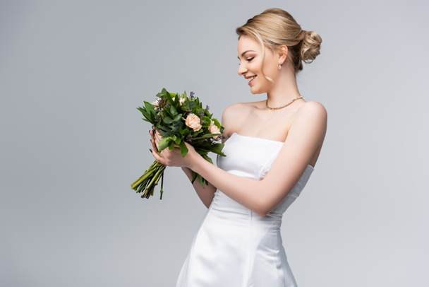 young and happy bride in white dress looking at bouquet of flowers isolated on grey  - Foto, Imagen