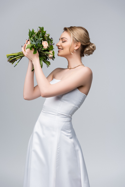 young and cheerful bride in white dress smelling bouquet of flowers isolated on grey  - Photo, Image