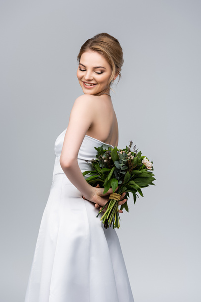 young and cheerful woman in white wedding dress holding bouquet of flowers isolated on grey  - Фото, зображення