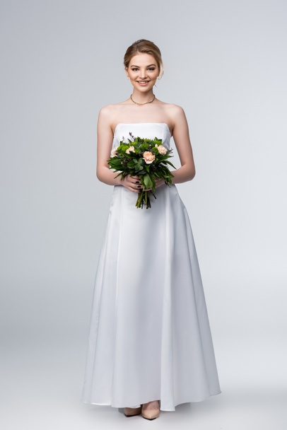 Lächelndes Mädchen im weißen Hochzeitskleid mit Blumenstrauß auf grau  - Foto, Bild