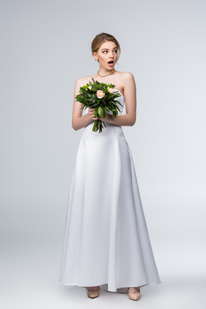 shocked girl in white wedding dress holding bouquet of flowers isolated on grey  - Foto, afbeelding