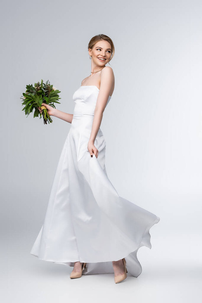 uśmiechnięta panna młoda dotykająca białej sukni ślubnej i trzymająca bukiet kwiatów na szarości  - Zdjęcie, obraz