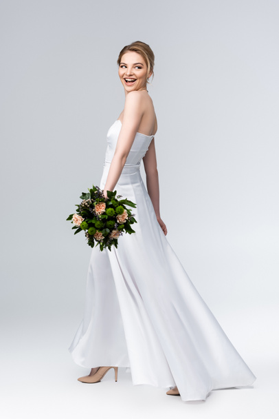 mariée attrayante dans une élégante robe de mariée tenant bouquet de fleurs et souriant sur gris - Photo, image