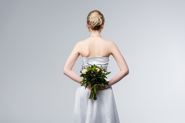 Rückansicht der Braut im eleganten Brautkleid mit Blumenstrauß hinter dem Rücken isoliert auf grau - Foto, Bild