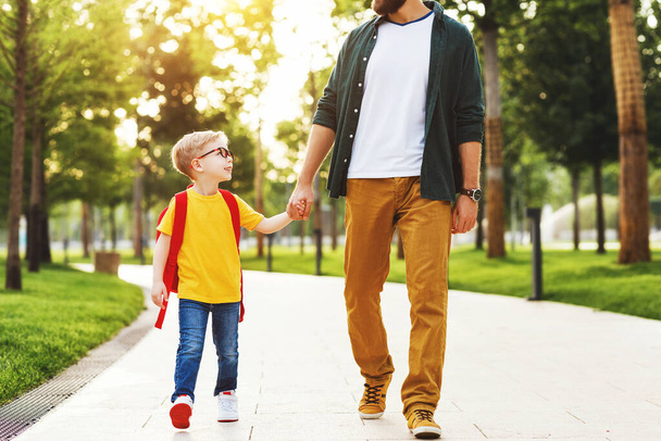 Onnellinen fiksu poika muistikirjat hymyillen ja katsellen sadon isä kävellessään kouluun vihreässä puistossa yhdessä - Valokuva, kuva