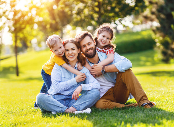 Cheerful boy and girl embracing happy parents from behind and looking at camera while resting on green grass on sunny day in par - Photo, Image