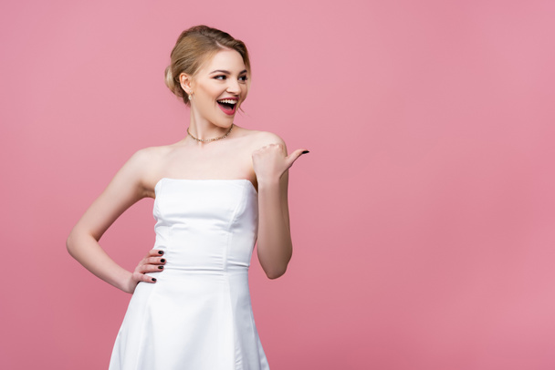 excited bride in white wedding dress pointing with thumb isolated on pink  - Fotografie, Obrázek