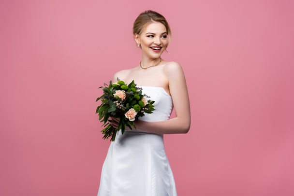 usmívající se nevěsta v bílých svatebních šatech drží květiny izolované na růžové  - Fotografie, Obrázek