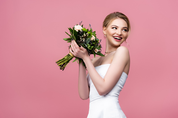 cheerful girl in white wedding dress holding flowers and looking away isolated on pink  - Foto, immagini
