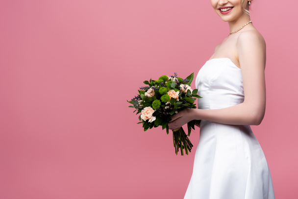 abgeschnittene Ansicht einer glücklichen Braut im weißen Hochzeitskleid mit Blumen auf rosa  - Foto, Bild