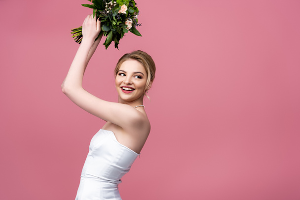 glückliche Braut im weißen Hochzeitskleid mit Blumen über dem Kopf isoliert auf rosa  - Foto, Bild
