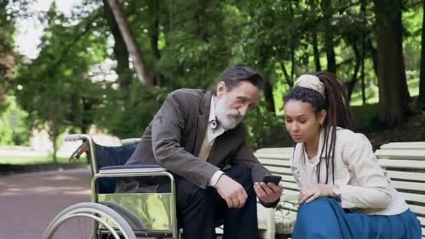 Family concept where esteemed sedentary old bearded man in wheelchair showing something his attractive modern confident young granddaughter with dreadlocks on mobile in green park - Πλάνα, βίντεο