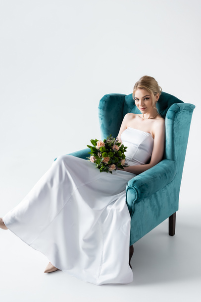 attractive bride in elegant wedding dress holding flowers while sitting in armchair on white - Φωτογραφία, εικόνα
