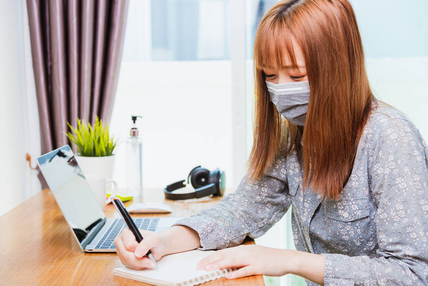 Asian young business woman wearing face mask protective working from home office with laptop computer he quarantines disease coronavirus or COVID-19 and write note booklist work today - Fotografie, Obrázek