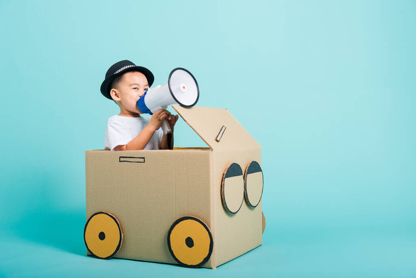 Happy Asian children boy smile in driving play car creative by a cardboard box imagination with megaphone, summer holiday travel concept, studio shot on blue background with copy space for text - Foto, Bild
