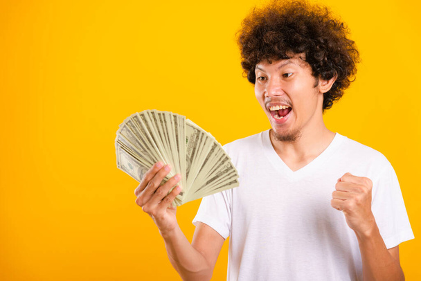 Asian handsome man with curly hair holding fans of money dollar bills isolate on yellow background with copy space for text - Fotó, kép