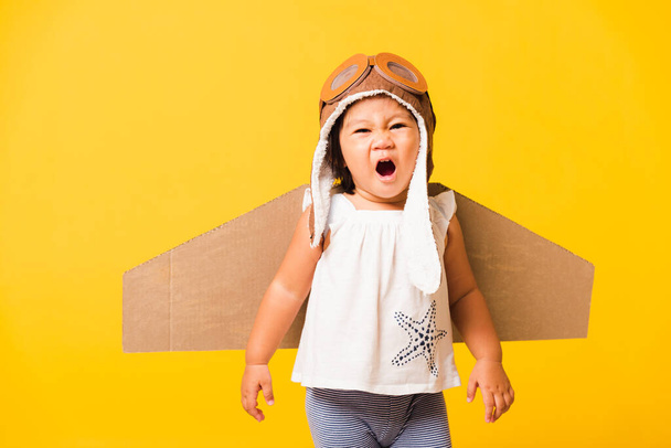 Happy Asian beautiful funny baby little girl smile wear pilot hat playing and goggles with toy cardboard airplane wings flying, studio shot isolated yellow background, Startup freedom concept - Foto, Imagen