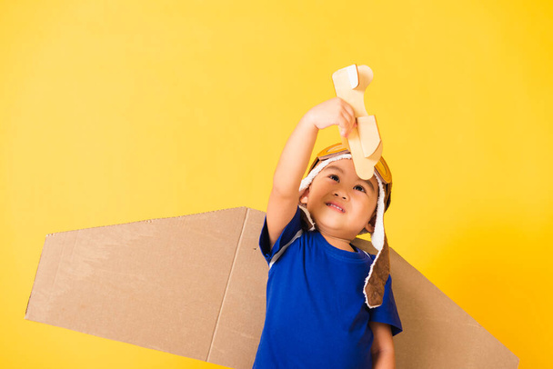 Happy Asian handsome funny child or kid little boy smile wear pilot hat play and goggles with toy cardboard airplane wings fly hold plane toy, studio shot isolated yellow background, Startup freedom - 写真・画像