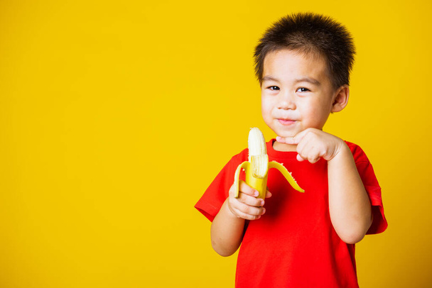 Happy portrait Asian child or kid cute little boy attractive smile wearing red t-shirt playing holds peeled banana for eating, studio shot isolated on yellow background - Фото, изображение