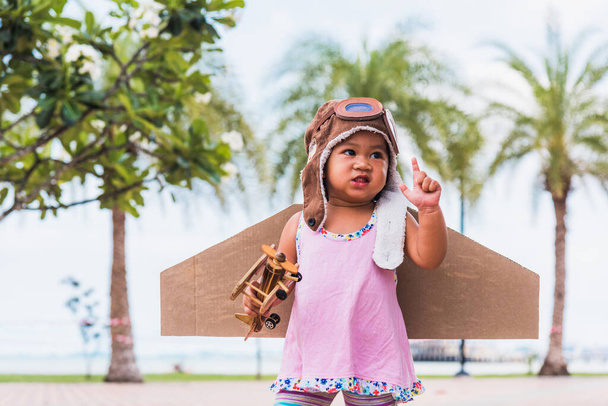 Happy Asian funny child or kid little girl smile wear pilot hat and goggles play toy cardboard airplane wing flying against summer sky cloud on trees garden background, Startup freedom concept - Фото, зображення