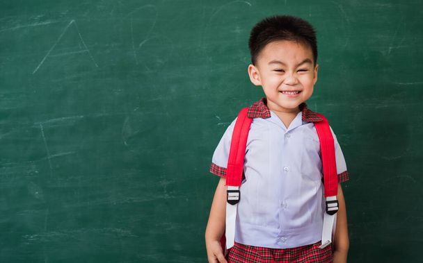Back to School. Happy Asian funny cute little child boy from kindergarten in student uniform with school bag stand smiling on green school blackboard, First time to school education concept - Fotografie, Obrázek