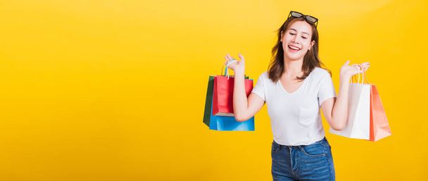 Asian Thai portrait happy beautiful cute young woman smiling stand with sunglasses excited holding shopping bags multi color looking down, studio shot isolated yellow background with copy space - Foto, afbeelding