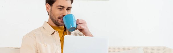 Plan panoramique d'un bel homme buvant du café et utilisant un ordinateur portable à la maison  - Photo, image