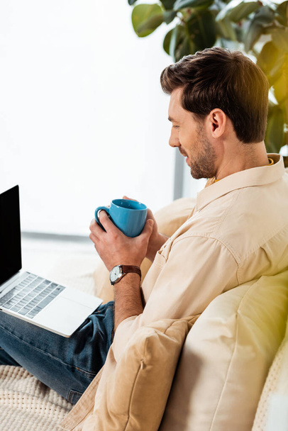 Vue latérale de l'homme tenant une tasse de café et regardant l'ordinateur portable sur le canapé  - Photo, image