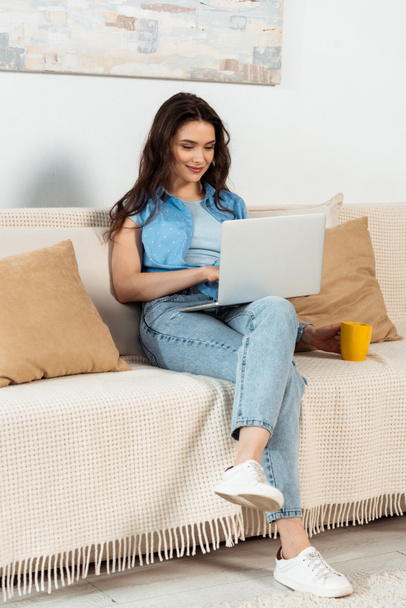 Beautiful woman using laptop and holding cup of coffee on couch  - Φωτογραφία, εικόνα