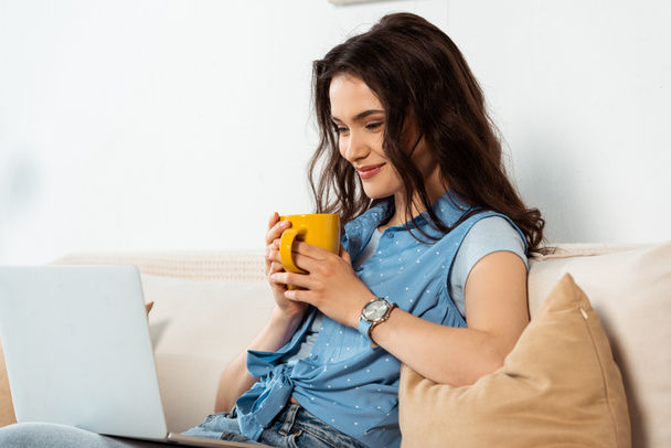 Sonriente freelancer sosteniendo la taza y mirando el portátil en el sofá
  - Foto, Imagen