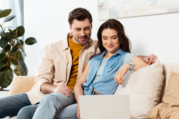 Selective focus of handsome man sitting near smiling girlfriend and laptop on couch  - Foto, Imagen