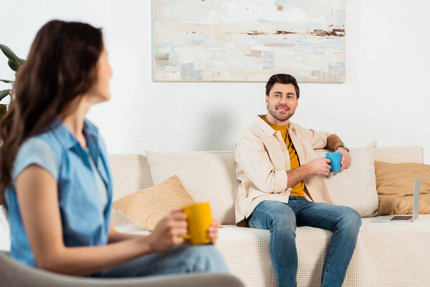 Selective focus of man holding cup of coffee near laptop and smiling at girlfriend in living room - Photo, Image