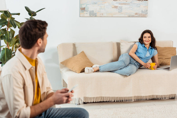 Selektiver Fokus der Frau, die Tasse neben Laptop auf Couch hält und Freund im Wohnzimmer anlächelt - Foto, Bild