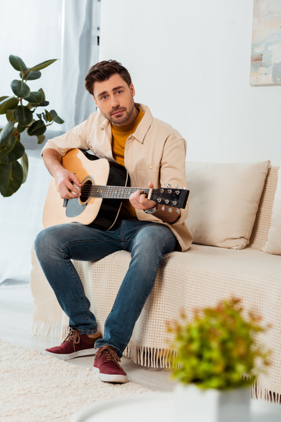 Selective focus of man looking at camera while playing acoustic guitar at home  - 写真・画像