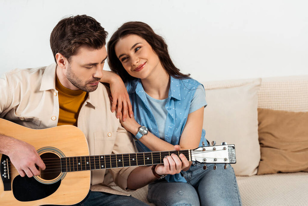 Smiling woman hugging and looking at boyfriend performing on acoustic guitar at home  - Foto, Imagem