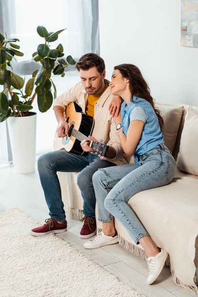 Smiling girl looking at boyfriend playing acoustic guitar at home  - Photo, Image